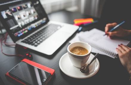 Mesa de trabalho com laptop, celular, caderno e um café servido na xícara.